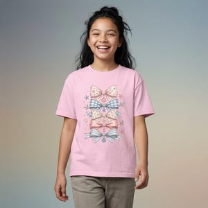 Puede incluir: Una camiseta rosa claro con un diseño de lazos decorativos en varios colores pastel. Los lazos incluyen patrones de estrellas, cuadros y rayas. El diseño se acentúa con pequeñas estrellas y elementos florales.