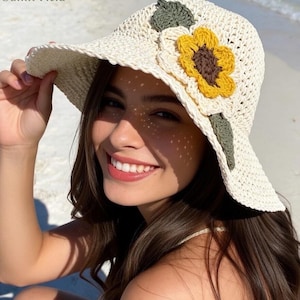 May include: A cream-colored, crocheted sun hat with a wide brim, adorned with a large yellow and brown sunflower and green leaves. The hat is labeled "Sunlit Field" in the upper left corner. The hat is being held up by a person.