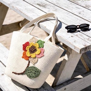 May include: A cream-colored woven tote bag featuring a large, multi-colored crocheted flower appliqué on the front. The flower has a dark brown center, yellow petals, and additional petals in red, orange, pink, and green. A brown crocheted stem and green leaf extend downward.