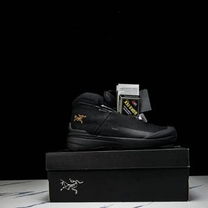 May include: Black hiking boot with a mesh upper, sitting on a black box. The boot has a high-top design, a black sole, and a gold logo on the side. A tag that says GORE-TEX is attached to the boot.