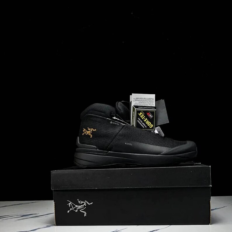 May include: Black hiking boot with a mesh upper, sitting on a black box. The boot has a high-top design, a black sole, and a gold logo on the side. A tag that says GORE-TEX is attached to the boot.
