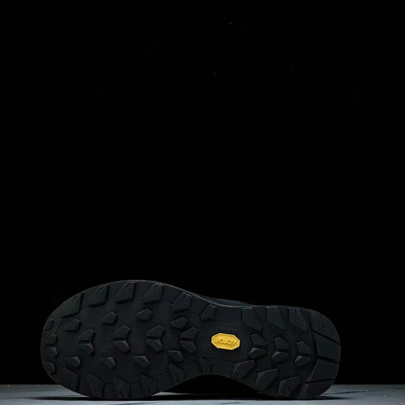May include: Black hiking boot sole with a textured pattern and a yellow Vibram logo. The sole is designed for traction and durability, suitable for outdoor activities. The boot is likely for hiking or trail running.