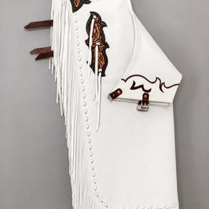 Puede incluir: Chaps de cuero blanco con largos flecos blancos en el lateral, detalles en cuero marrón con grabado floral en la parte superior y central, y correas de cuero marrón con hebillas metálicas para ajustar alrededor de la pierna.