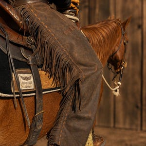 May include: Dark brown leather chaps featuring long fringe detailing along the outer legs, worn over trousers while seated on a horse. The chaps are secured with a wide leather belt and include a saddle pad with a patch reading Relentless Cactus.