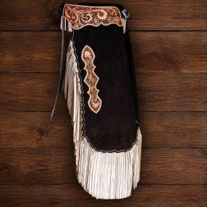 May include: A pair of black suede chaps featuring a tan leather yoke and a vertical side panel, both decorated with intricate floral tooling. The garment is finished with long, cream-colored leather fringe along the outer edge and a metal buckle.