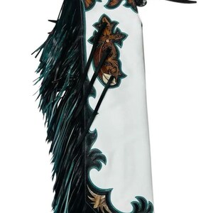 Peut inclure: Chaps en cuir blanc avec des accents en cuir brun à motifs floraux, une bordure noire et un passepoil turquoise. Le panneau latéral présente une croix en cuir brun, tandis qu'une longue frange noire et turquoise pend sur le bord extérieur.