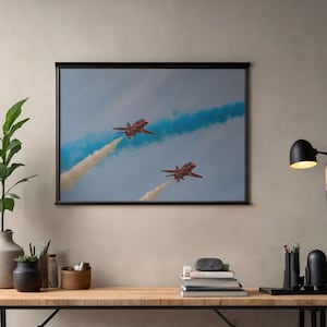 May include: A framed print showing two red and white jet aircraft in flight, with white and coloured smoke trails against a blue sky. The jets are angled upwards, creating a dynamic composition. The print is displayed in a black frame.