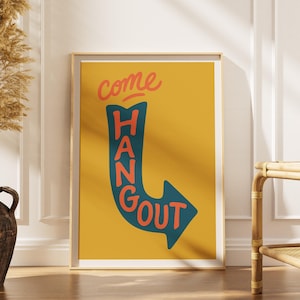 May include: A rectangular framed wall art print featuring a mustard yellow background. A dark teal arrow shape curves downward, containing the orange text "COME HANG OUT" written in a bold, sans-serif font. The print is displayed in a light wood frame.