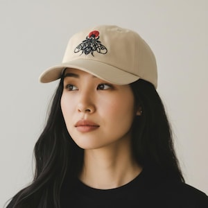 Peut inclure: Casquette de baseball beige avec un motif de mouche brodé en noir et rouge. La casquette a une visière incurvée et une construction classique à six panneaux. Le motif de la mouche présente des détails complexes et un point rouge au-dessus.