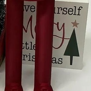 May include: A deep red, 3D-printed decorative object shaped like a pair of legs wearing boots, with a cylindrical top featuring a molded belt and buckle detail. The structure is solid red and stands upright on two feet, resembling a festive character.