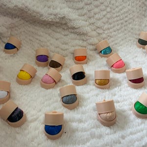 May include: A collection of small, cylindrical beige containers, each holding a distinctively colored fabric insert. The inserts feature a wide variety of solid and patterned colors, including blue, yellow, pink, black, green, orange, and metallic tones.