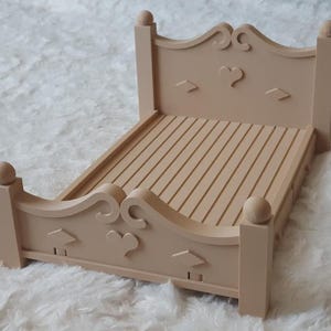 May include: A light beige doll bed featuring a headboard and footboard with carved heart and diamond shapes, curved top edges, and spherical finials on each corner post. The bed base consists of a series of parallel horizontal slats.