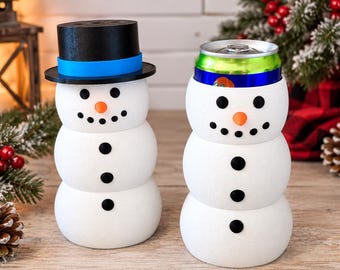 May include: Two white snowman-shaped beverage insulators. The left one wears a black top hat with a blue band. The right one holds a metal soda can with a green and blue label. Both feature black button eyes, a carrot-colored nose, and black buttons.