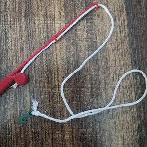 Könnte beinhalten: Eine rote Spielzeug-Angelrute aus Kunststoff mit einer weißen Schnur an der Spitze und einem kleinen roten Rollenelement am Griff. Ein kleiner grüner Kunststoffhaken ist am Ende der weißen Schnur befestigt, die in einer losen Schlaufe hängt.