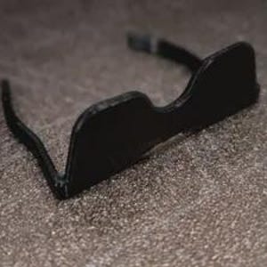 May include: A pair of black, solid-frame glasses featuring a distinct, continuous bridge design that connects the two lens areas. The frames are matte black with a smooth finish, showcasing a curved top edge and thin, straight temples extending backward.