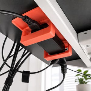 May include: A bright orange under-desk cable management mount holding a black power brick and power cord. The mount is attached to the underside of a black desk surface, with additional black cables bundled together with a hook-and-loop strap.