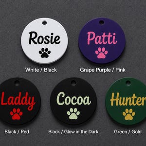 May include: Five circular pet identification tags with a hole at the top. The tags feature the names Rosie, Patti, Laddy, Cocoa, and Hunter in various colors, each accompanied by a paw print graphic. The color combinations are white/black, grape purple/pink, black/red, black/glow-in-the-dark, and green/gold.