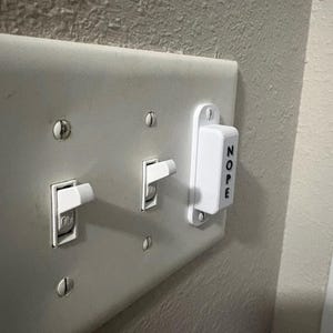 May include: A white plastic light switch cover plate featuring two standard toggle switches in the on position and a rectangular white plastic block attached to the right side with the word NOPE printed vertically in black capital letters.