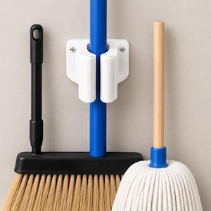 May include: A white plastic wall-mounted mop and broom holder with two spring-loaded clips. The holder secures a blue handle, flanked by a black broom with tan bristles on the left and a white cotton string mop with a light wood handle on the right.