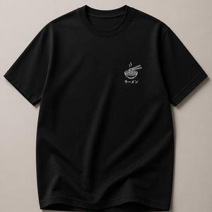 May include: Black t-shirt featuring a white graphic of a ramen bowl with chopsticks and steam. The Japanese word "ラーメン" is printed below the bowl. The shirt has a crew neck and short sleeves.