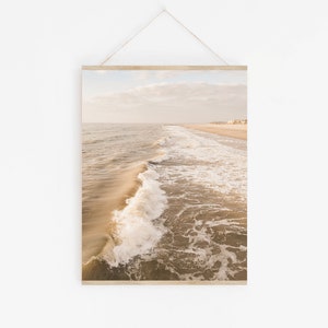 Pu&ograve; includere: Una stampa incorniciata di una scena di spiaggia. La fotografia cattura le onde dell'oceano che si infrangono sulla riva. L'acqua &egrave; di colore marrone chiaro e il cielo &egrave; azzurro pallido. La stampa &egrave; appesa da una barra di legno e una corda.
