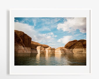 May include: A photograph of a scenic landscape featuring a body of water with red rock formations in the background. The sky is blue with white clouds. The water is calm and reflects the sky and clouds.