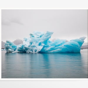 May include: A framed print of a vibrant blue iceberg floating in calm, blue water. The iceberg has a textured surface and is set against a backdrop of a cloudy sky and distant mountains. The overall scene evokes a sense of tranquility.