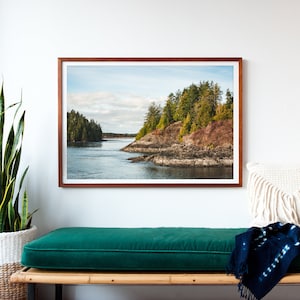 May include: A framed landscape photograph depicts a coastal scene with a body of water and forested islands. The photograph is in a brown wooden frame. The water is a blue-gray color, and the sky is cloudy. The trees are green and brown.
