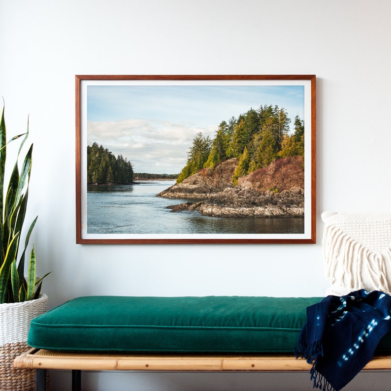 May include: A framed landscape photograph depicts a coastal scene with a body of water and forested islands. The photograph is in a brown wooden frame. The water is a blue-gray color, and the sky is cloudy. The trees are green and brown.