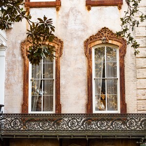 May include: Exterior view of a historic building with ornate, rust-colored window frames and a decorative wrought-iron balcony. The windows have sheer curtains, and the building's facade has a textured, aged appearance. Green foliage frames the top.