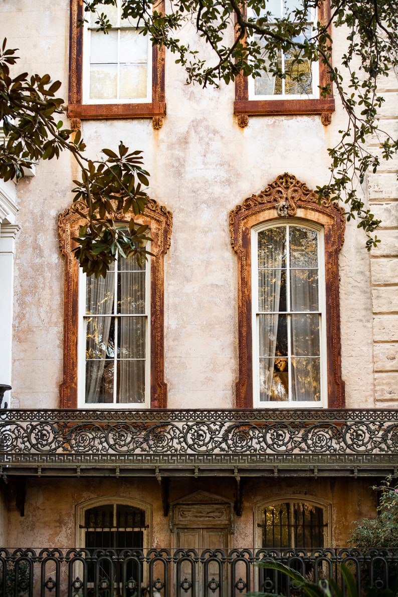 May include: Exterior view of a historic building with ornate, rust-colored window frames and a decorative wrought-iron balcony. The windows have sheer curtains, and the building's facade has a textured, aged appearance. Green foliage frames the top.
