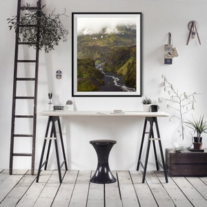 May include: A framed landscape photograph of a canyon with a river winding through it. The canyon walls are green with patches of snow, and the sky is overcast. The photograph is displayed on a white wall above a desk with a black stool.