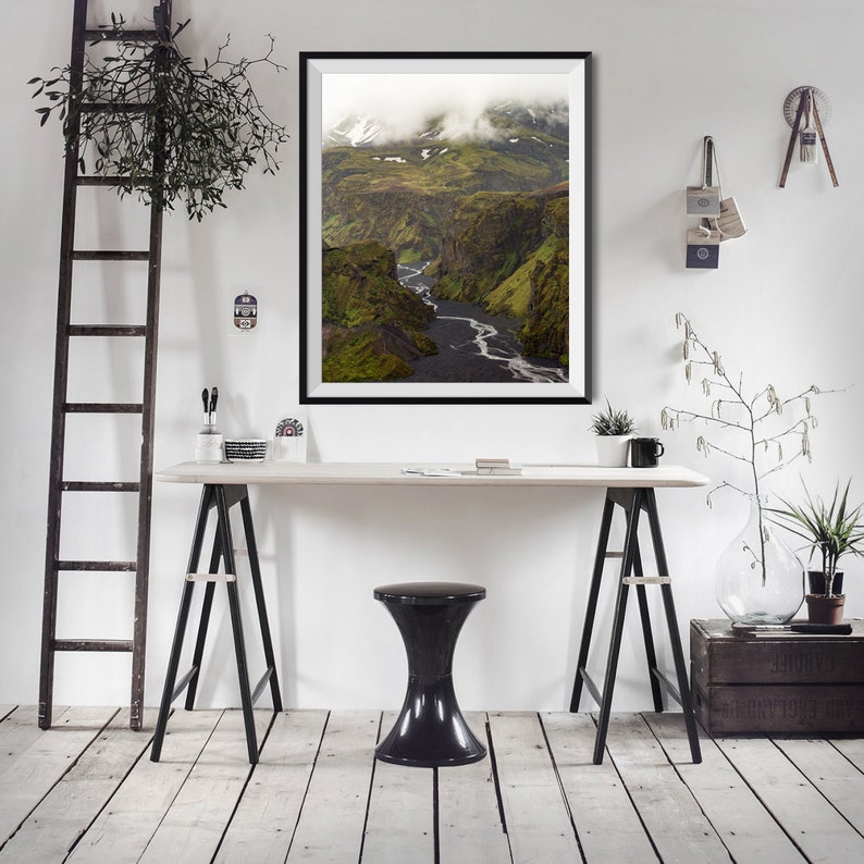 May include: A framed landscape photograph of a canyon with a river winding through it. The canyon walls are green with patches of snow, and the sky is overcast. The photograph is displayed on a white wall above a desk with a black stool.