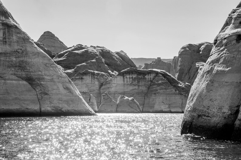 以下が含まれることがあります： 大きな岩石層のある湖の風景の白黒写真。水は太陽光の中で輝き、光を反射しています。岩石層は層状で質感があり、劇的な風景を作り出しています。