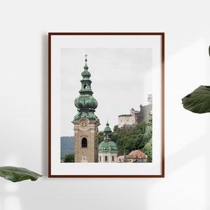 May include: Framed print of a European cityscape. The image shows a tall church tower with a green dome and spire, a castle on a hillside, and other buildings. The print is in a brown frame and is displayed against a white wall.