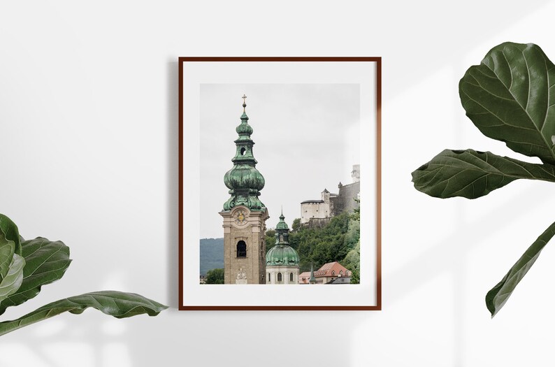 May include: Framed print of a European cityscape. The image shows a tall church tower with a green dome and spire, a castle on a hillside, and other buildings. The print is in a brown frame and is displayed against a white wall.