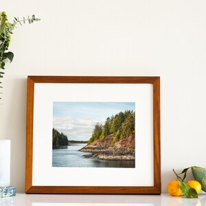 May include: A framed landscape photograph featuring a coastal scene with a body of water, rocky shoreline, and evergreen trees. The frame is made of wood. A vase with greenery, a book, a bowl, and oranges are also present.