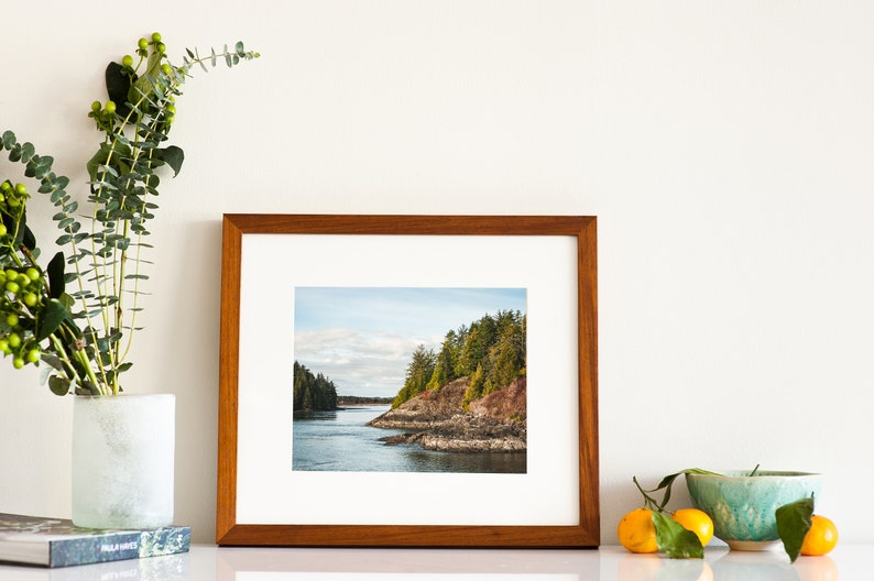 May include: A framed landscape photograph featuring a coastal scene with a body of water, rocky shoreline, and evergreen trees. The frame is made of wood. A vase with greenery, a book, a bowl, and oranges are also present.