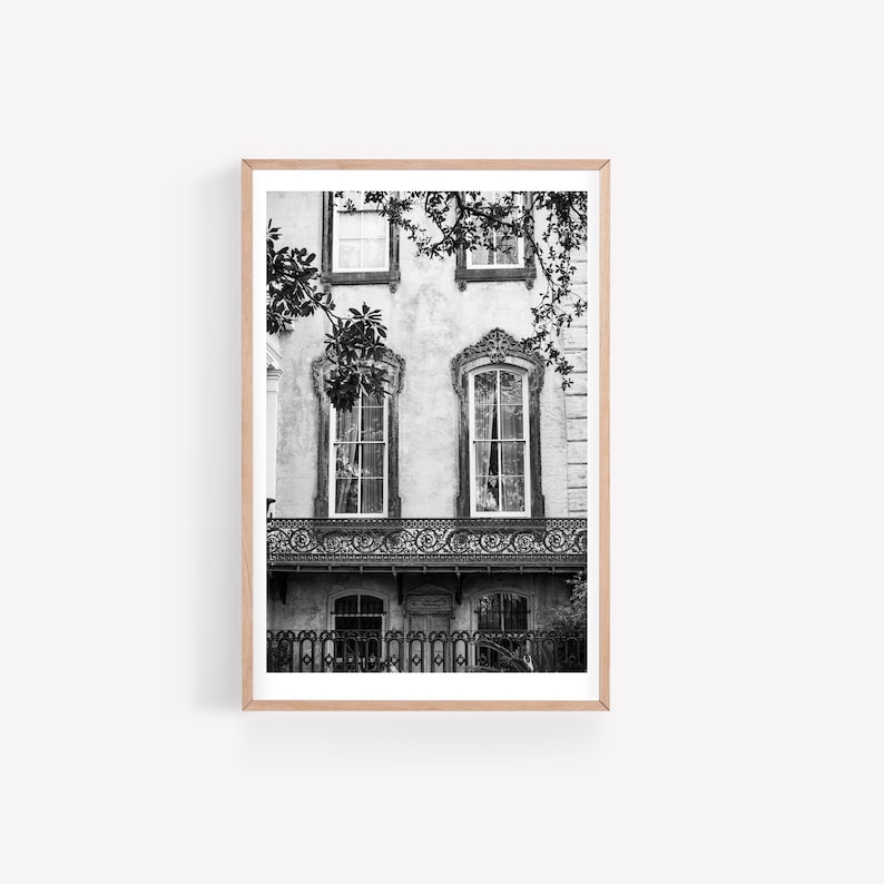 May include: Black and white photograph of a historic building facade. The image features ornate windows, a wrought-iron balcony, and a wooden frame. The building is partially obscured by tree branches, adding a natural element to the architectural scene.