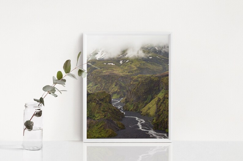 May include: A framed landscape photograph of a canyon with a river winding through it. The canyon walls are green with vegetation, and the riverbed is dark. The top of the canyon is covered in clouds.