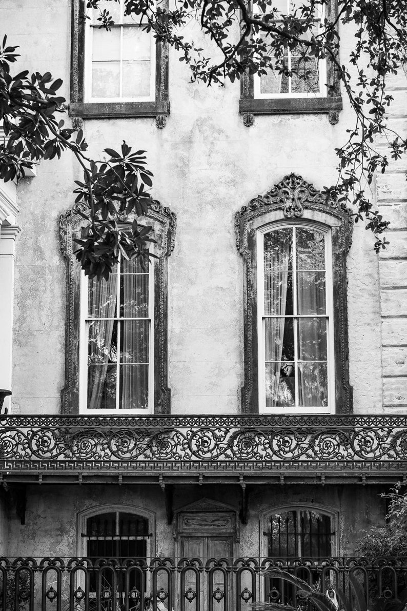 May include: Black and white photograph of a historic building facade. The image features ornate windows with decorative frames, a wrought-iron balcony, and a textured exterior wall. Foliage frames the top of the building.