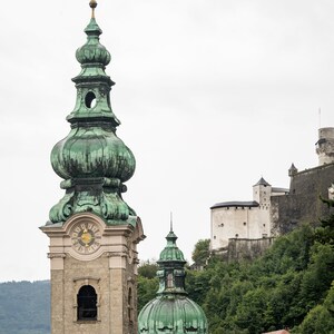 May include: A stone church tower with a green, onion-shaped dome and a clock face. A smaller dome and a castle on a hillside are visible in the background. The sky is overcast.