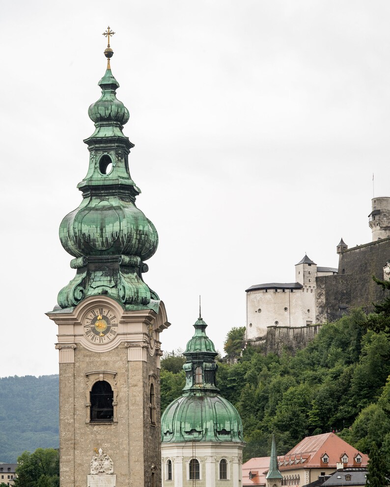 May include: A stone church tower with a green, onion-shaped dome and a clock face. A smaller dome and a castle on a hillside are visible in the background. The sky is overcast.