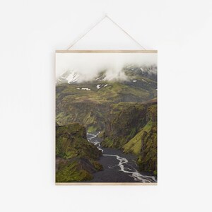 May include: A framed landscape print featuring a deep canyon with a river winding through it. The canyon walls are covered in green vegetation, and the sky is filled with clouds. The print is displayed on a white wall.
