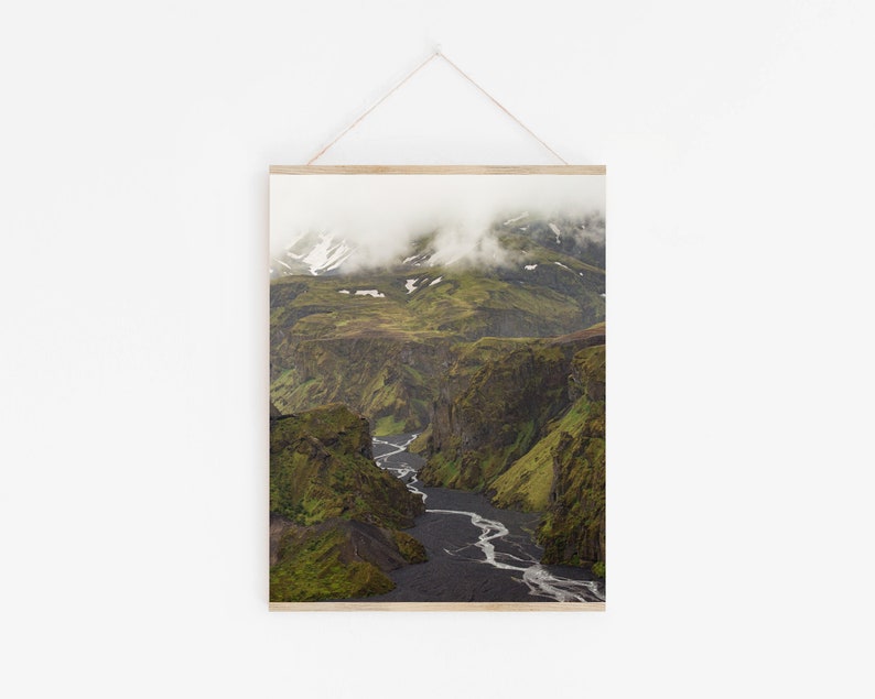 May include: A framed landscape print featuring a deep canyon with a river winding through it. The canyon walls are covered in green vegetation, and the sky is filled with clouds. The print is displayed on a white wall.