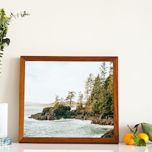 Pu&ograve; includere: Fotografia incorniciata di un paesaggio costiero roccioso con alberi sempreverdi. L'immagine &egrave; in una cornice di legno marrone. Un vaso con steli verdi e una ciotola con arance sono in primo piano.