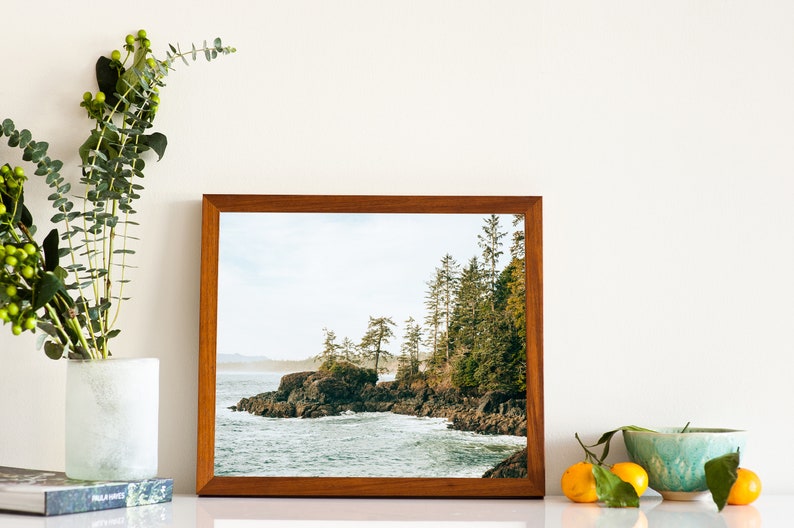 Pu&ograve; includere: Fotografia incorniciata di un paesaggio costiero roccioso con alberi sempreverdi. L'immagine &egrave; in una cornice di legno marrone. Un vaso con steli verdi e una ciotola con arance sono in primo piano.