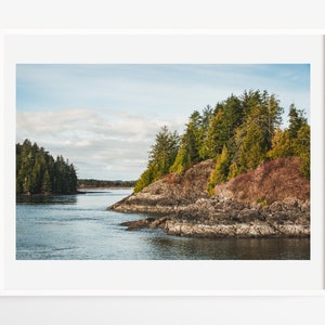 May include: A framed landscape print featuring a coastal scene. The image shows a body of water surrounded by rocky shores and lush green trees. The sky is a mix of blue and white, creating a serene and natural aesthetic.