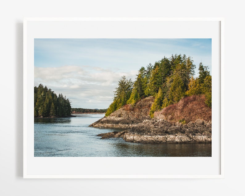 May include: A framed landscape print featuring a coastal scene. The image shows a body of water surrounded by rocky shores and lush green trees. The sky is a mix of blue and white, creating a serene and natural aesthetic.