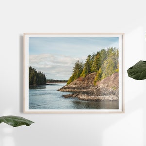 May include: A framed landscape print featuring a coastal scene. The image shows a body of water surrounded by rocky cliffs and lush green trees under a cloudy sky. The frame is a light wood color.